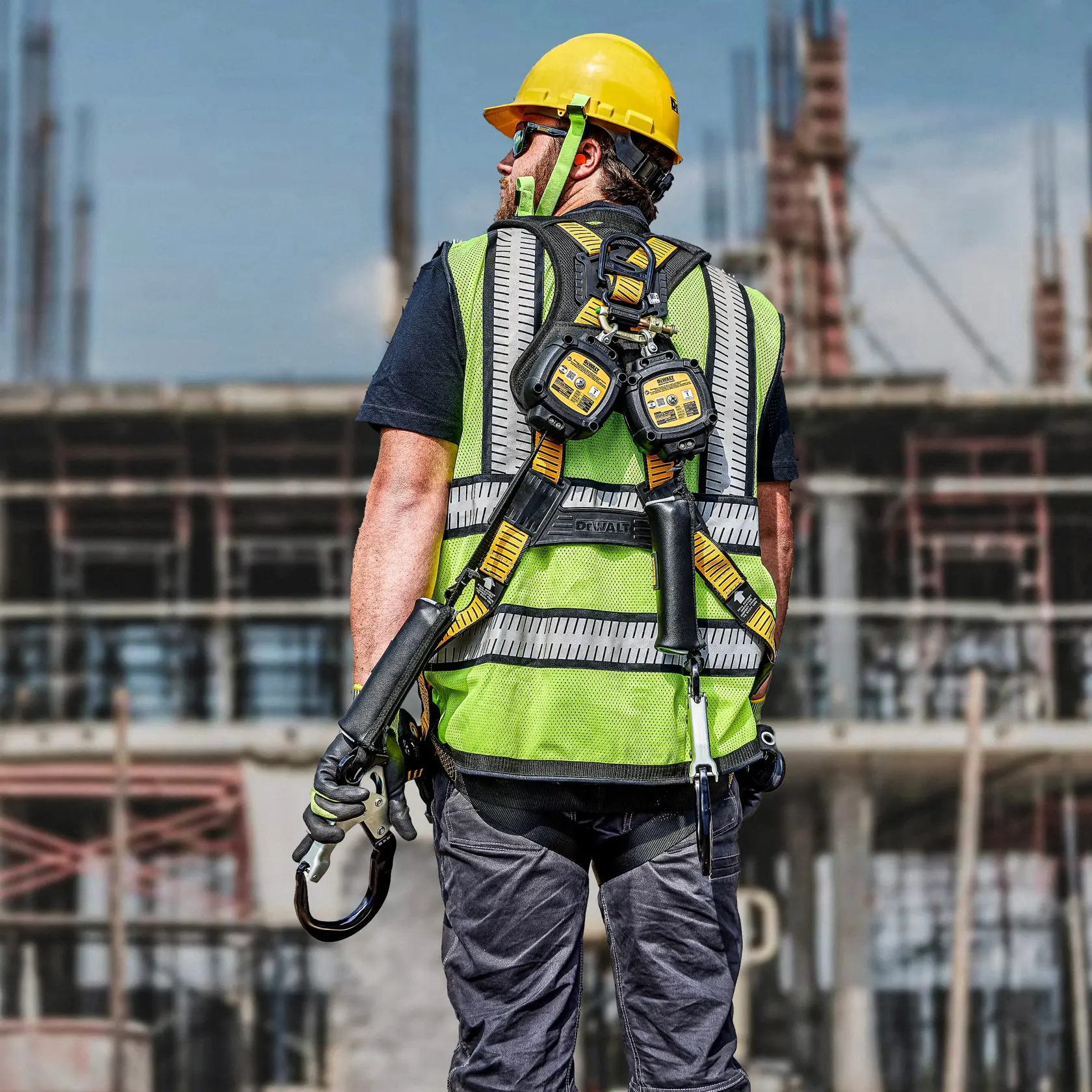 A construction worker wearing a yellow hard hat, a green high-visibility vest, and a DEWALT safety harness with lanyards, standing on a construction site. The worker’s face is blurred for privacy.