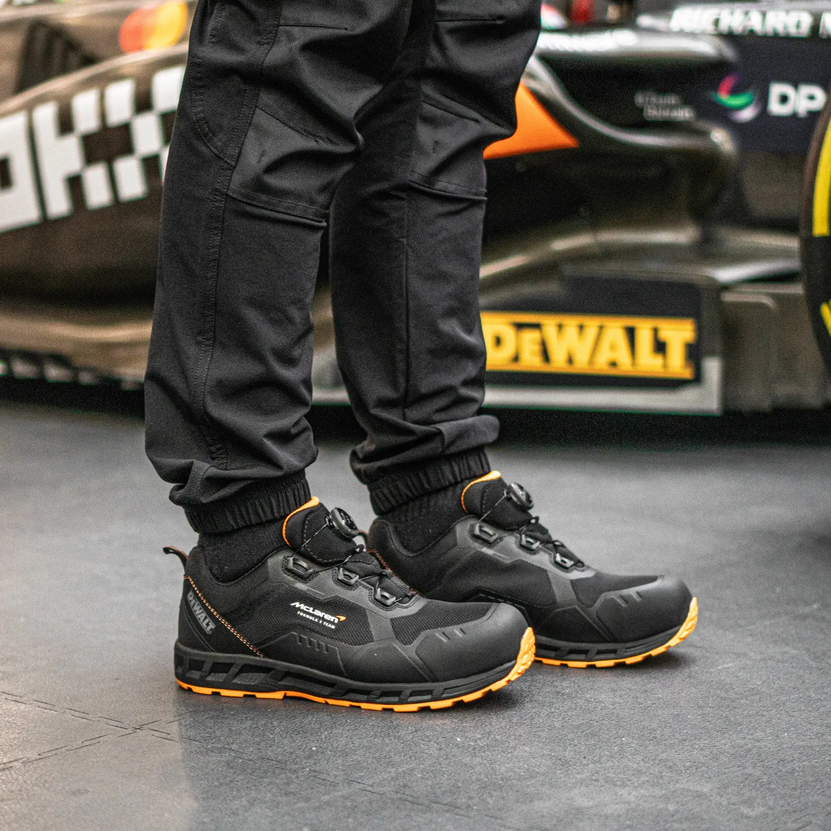 Side view facing right of the DEWALT x McLaren Torque Men's Safety Shoes in black on model on location with McLaren F1 car in background