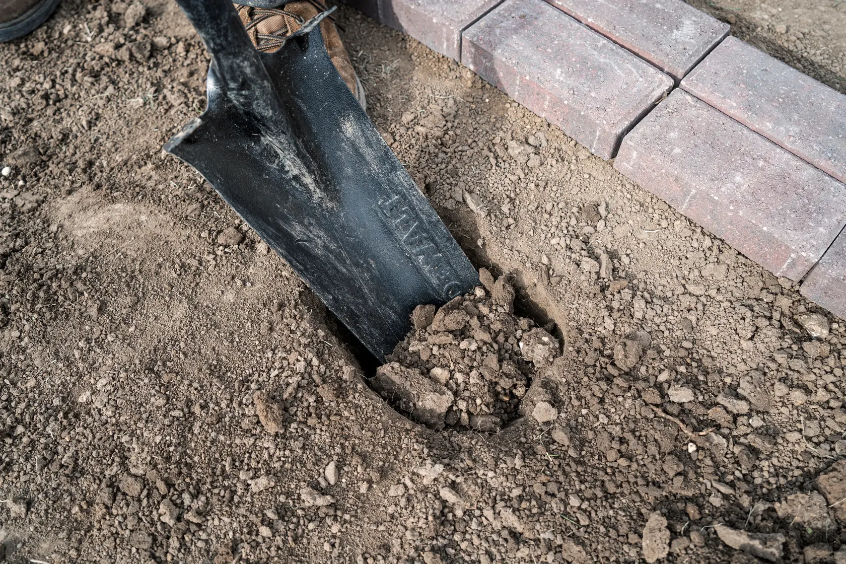 A close-up view of a shovel digging into soil next to a row of concrete pavers in an outdoor setting.