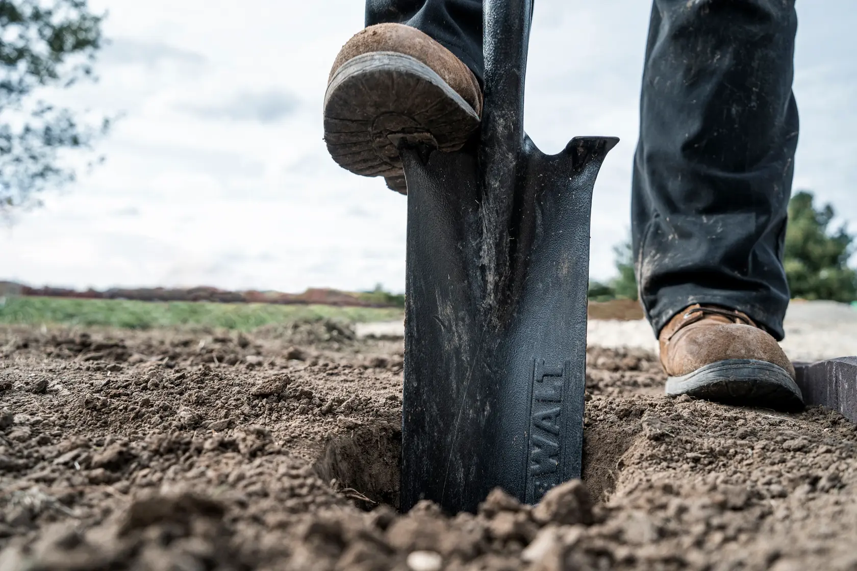 Close-up view of a person digging in soil with a DEWALT shovel. The person's boot is pressing down on the shovel blade, working in an outdoor, dirt area.
