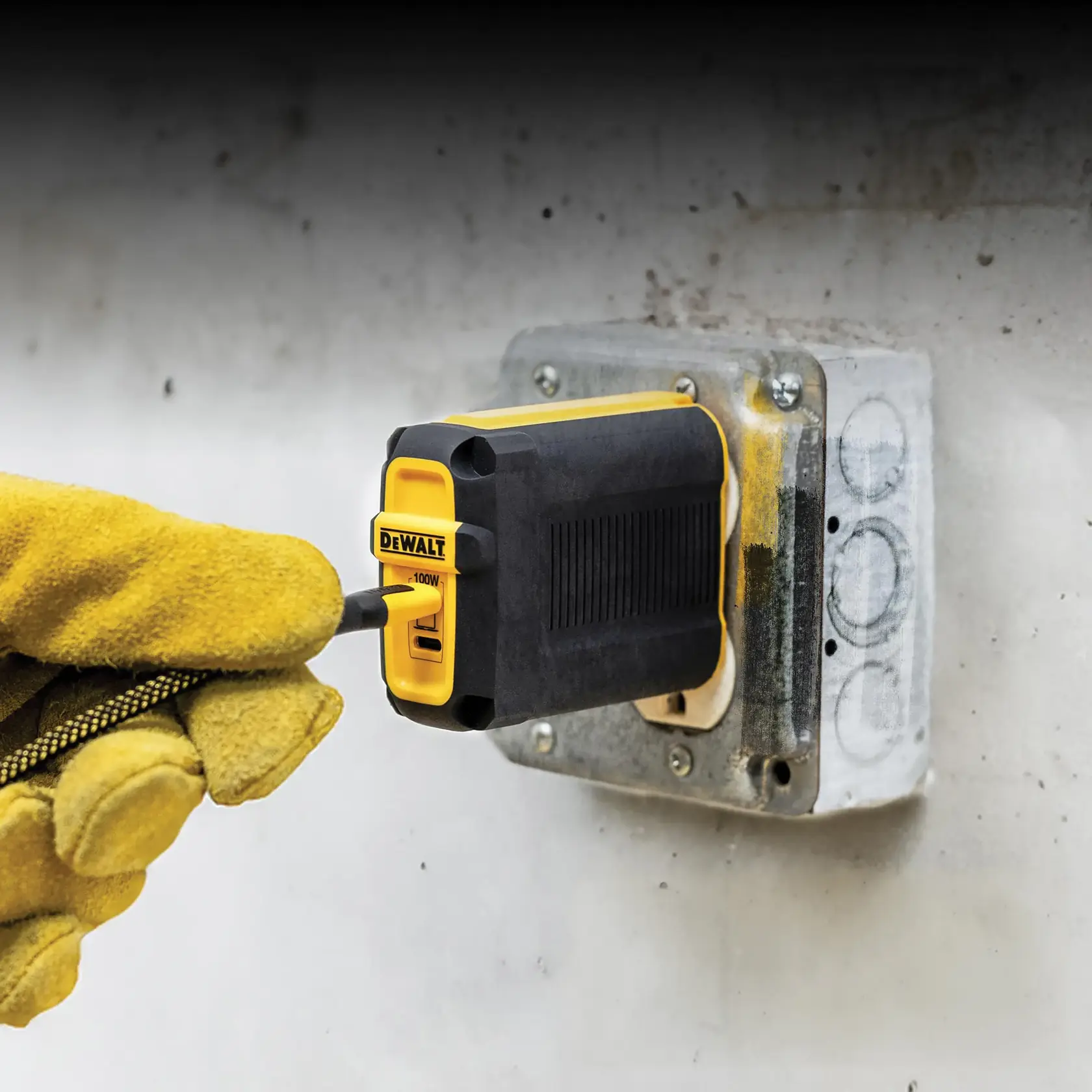 A person wearing a yellow work glove plugs a cable into a DEWALT power adapter that is connected to a wall outlet mounted on a metal electrical box.