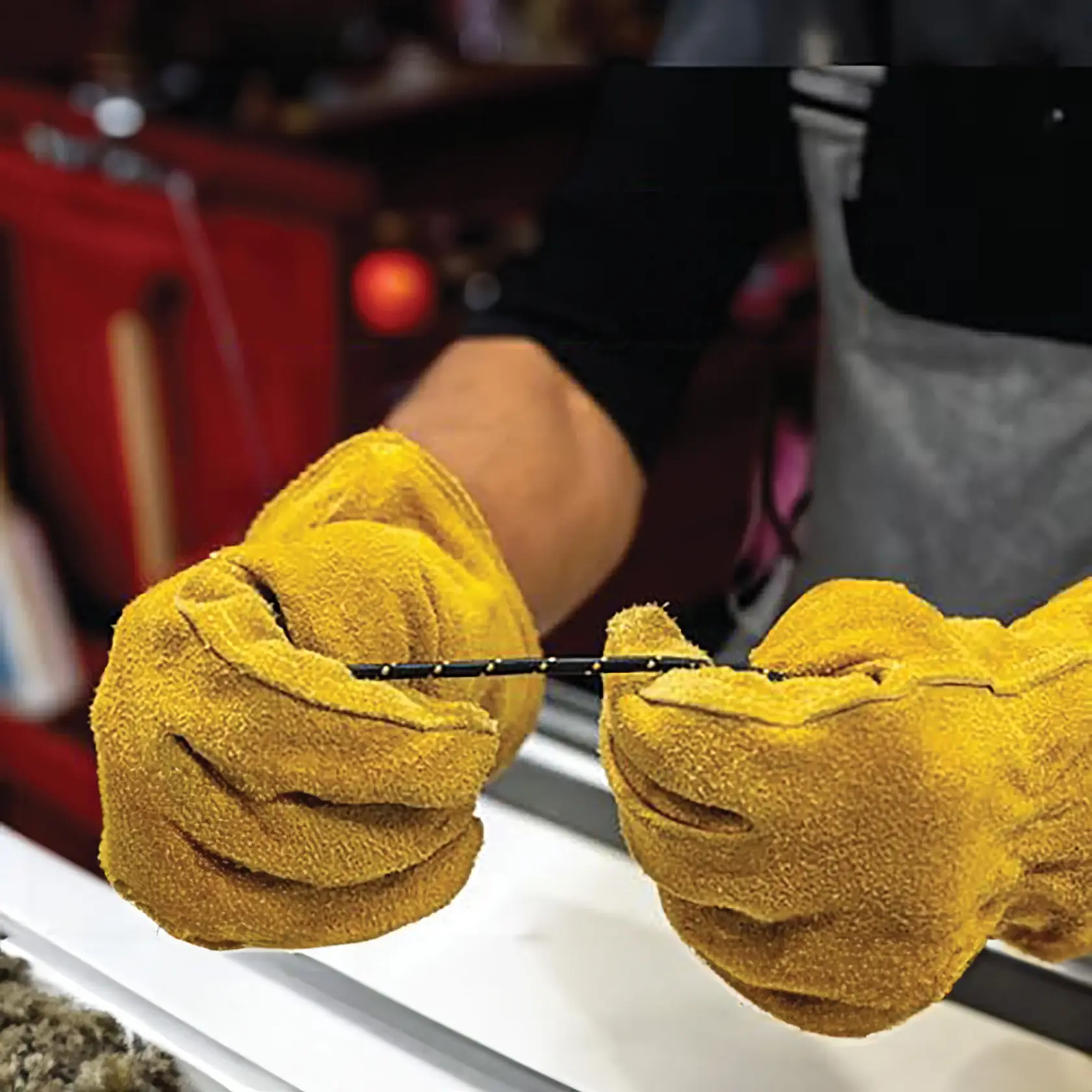 A person wearing yellow protective work gloves is holding a small black rod or tool, with a background of a workshop or workbench.