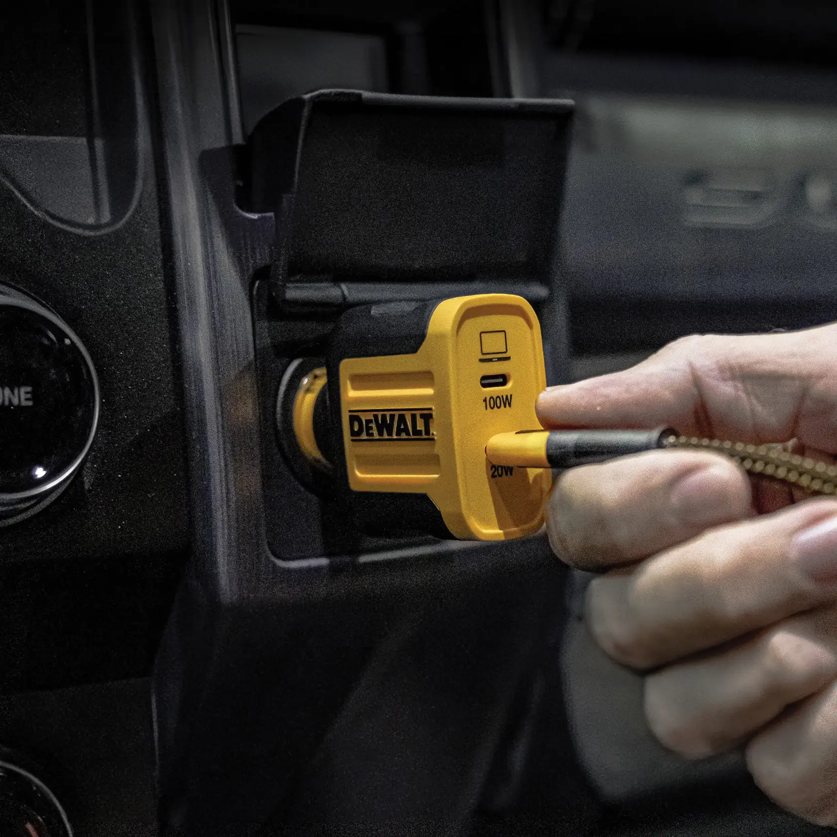 A DEWALT branded yellow and black car charger is plugged into a vehicle's power outlet. A hand is connecting a cable to the charger, which features a USB port and power indicators labeled 100W and 20W.