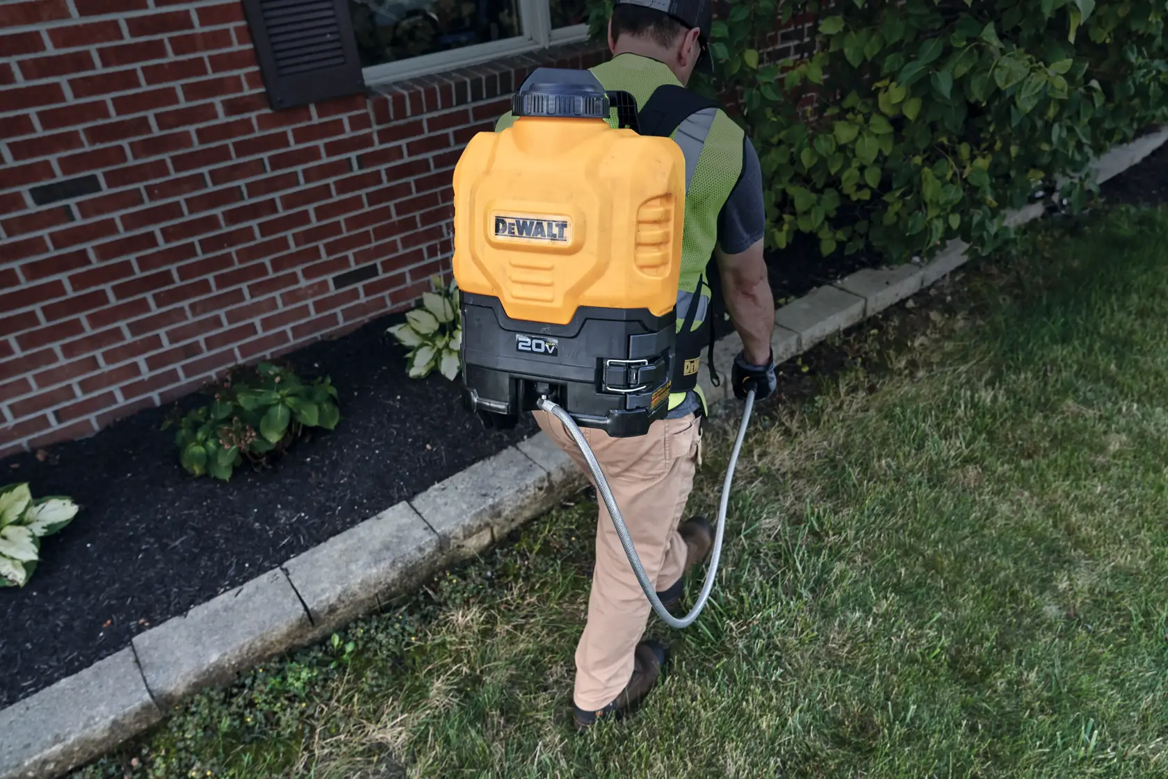 A person wearing a yellow and black DeWalt 20V backpack sprayer is working in a garden beside a brick house, spraying the lawn and landscaping.