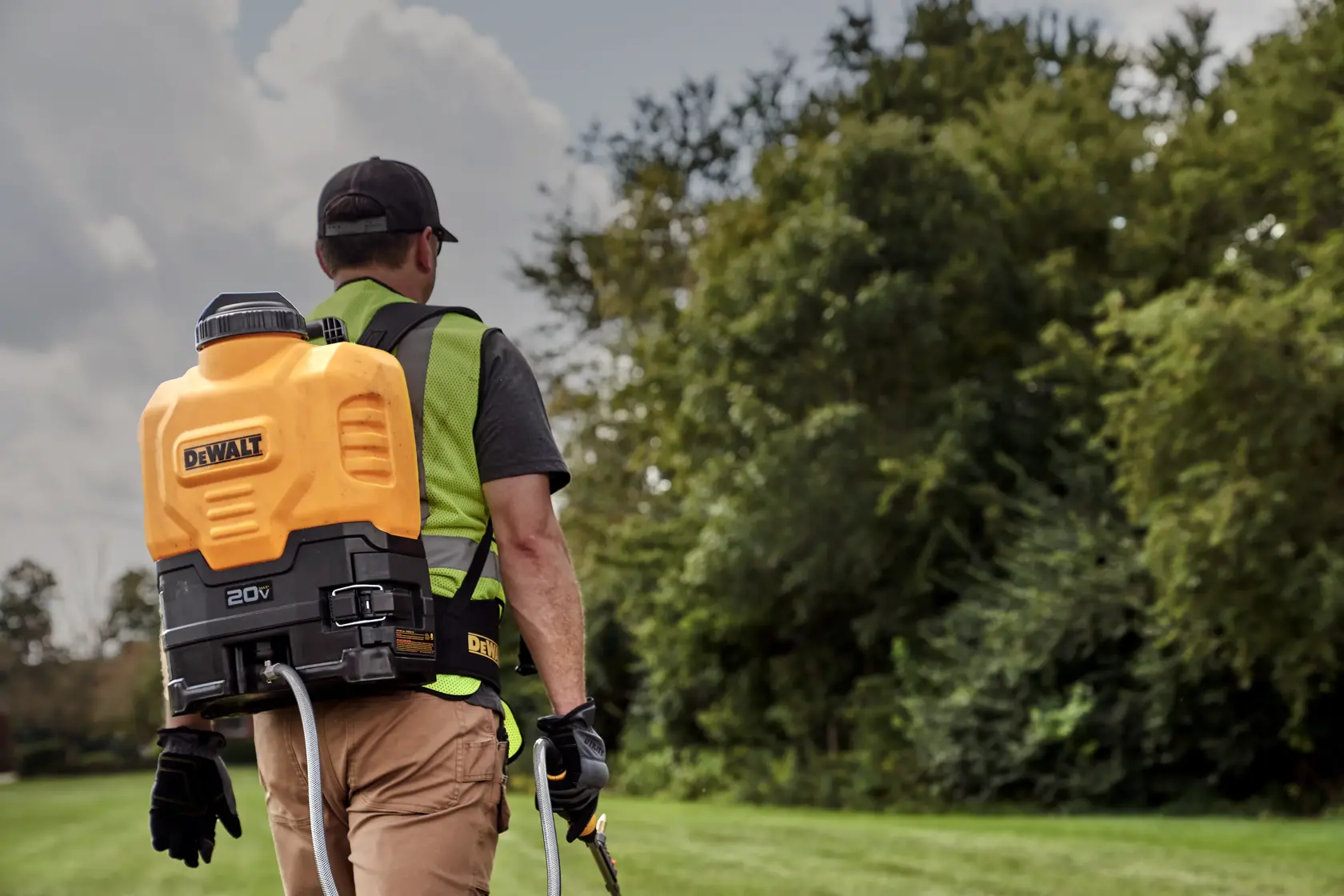 A person outdoors wearing a DEWALT 20V battery-powered backpack sprayer, walking on grass near trees.