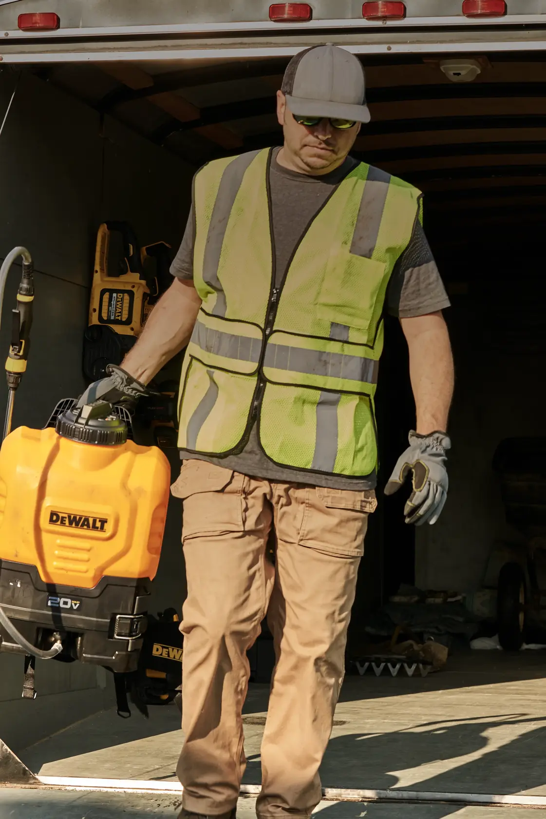 A person wearing a high-visibility safety vest, gloves, and a cap stands in a trailer while holding a yellow DeWalt 20V tool or equipment. Other tools can be seen mounted on the trailer walls.