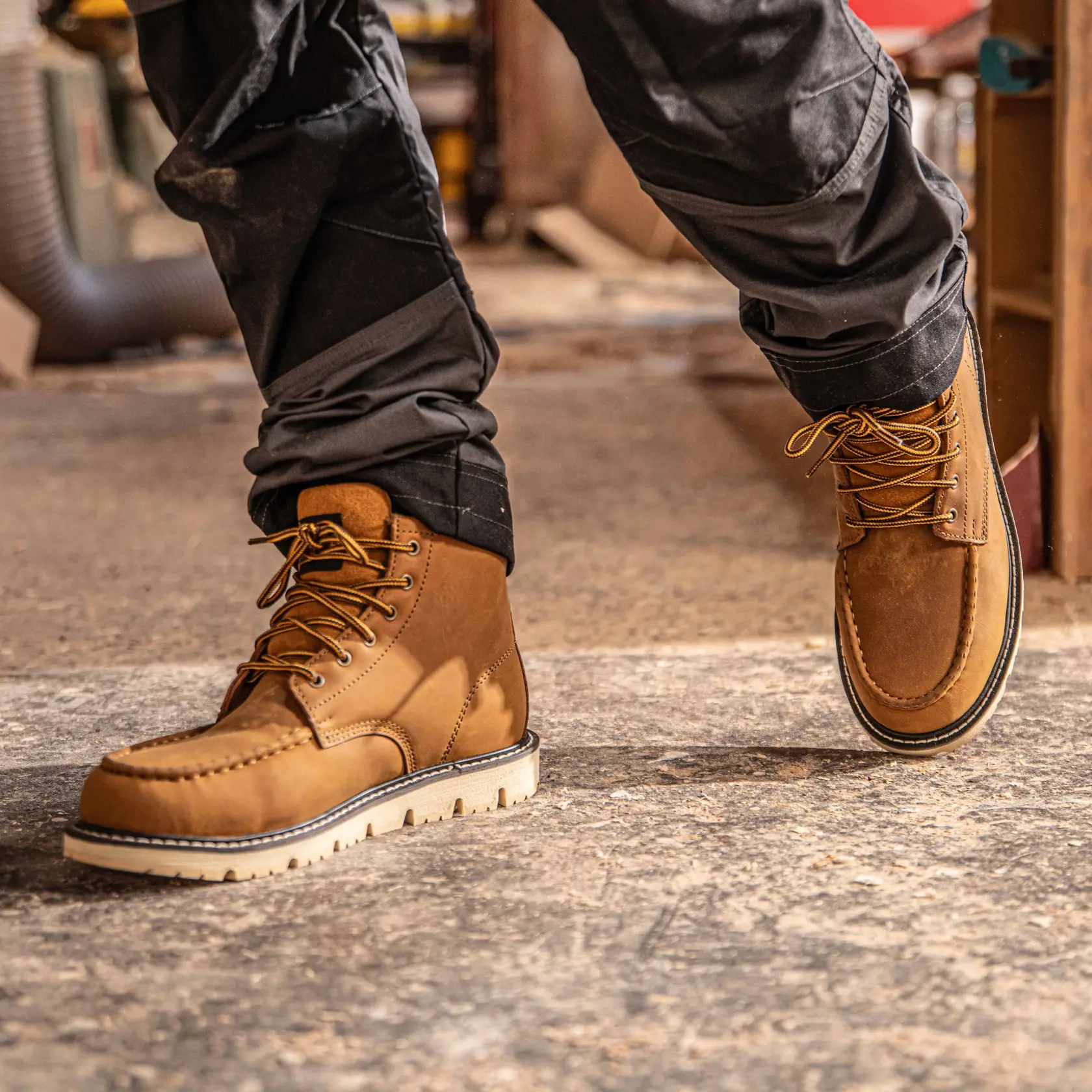 A close-up view of a person wearing tan DEWALT work boots with thick soles and yellow laces, standing indoors on a concrete floor.