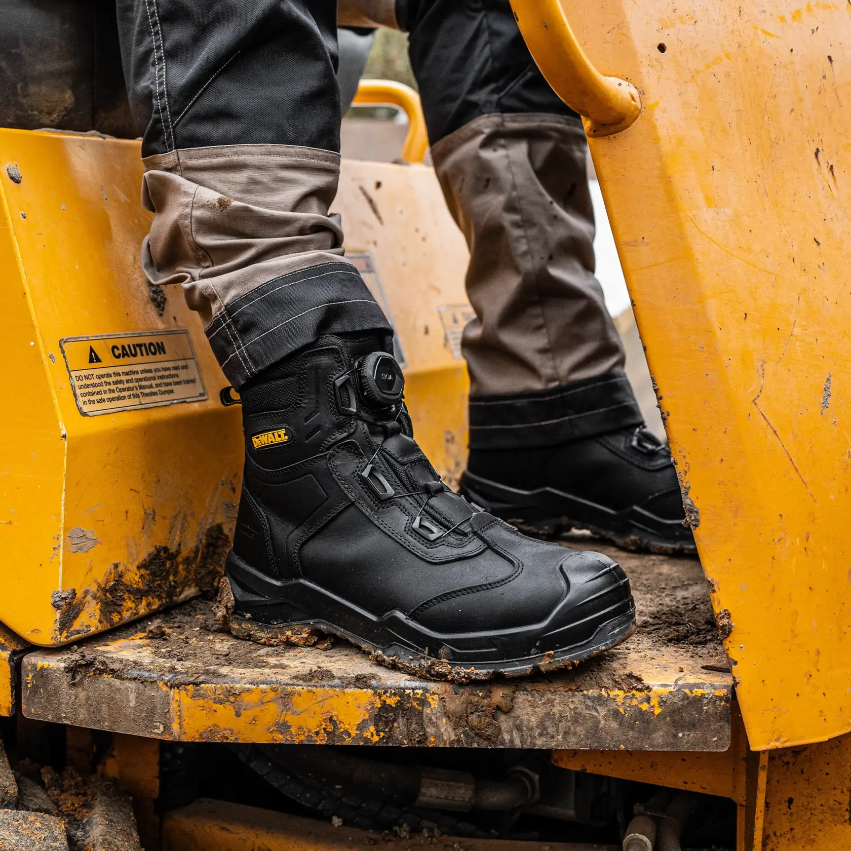 Close-up view of a person wearing black DEWALT work boots standing on a muddy, yellow construction vehicle. The boots are rugged and designed for outdoor work environments.