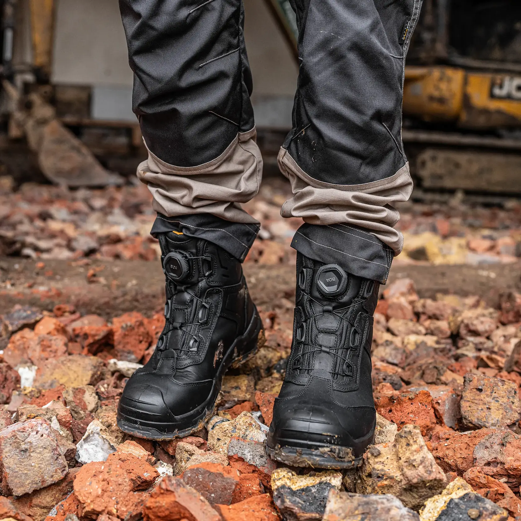 A pair of black DEWALT work boots with BOA lacing system worn by a person standing on rough, broken bricks and rocks at a construction site. The boots appear sturdy and are designed for protection and durability.
