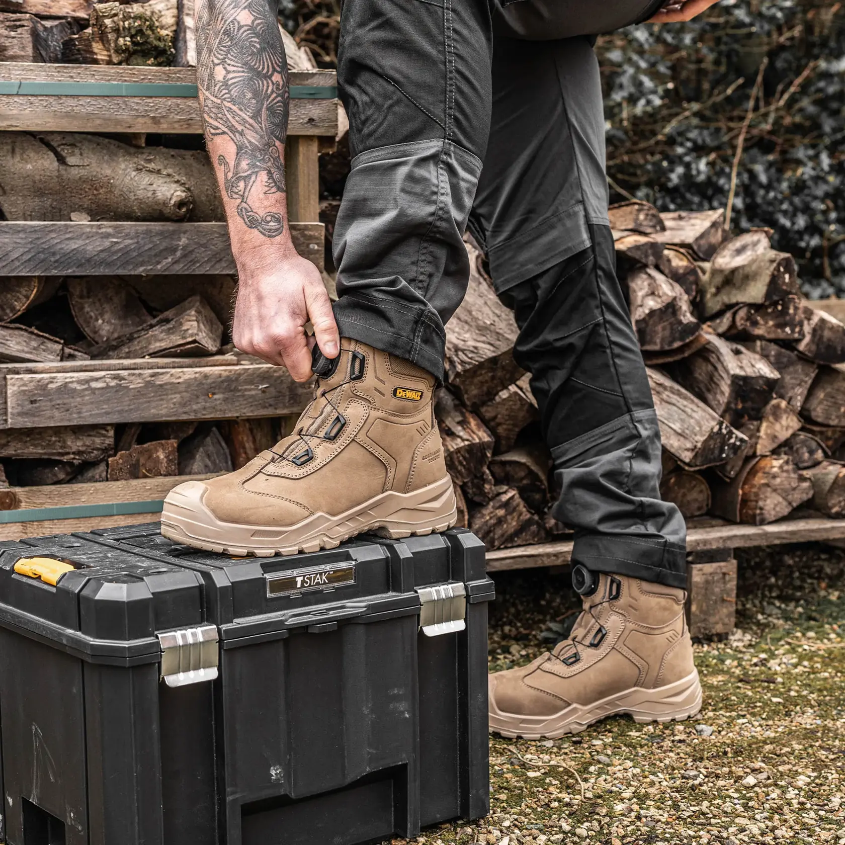 A person wearing tan DEWALT work boots adjusts the fit using a dial mechanism on the side of the boot. The person is standing outdoors next to a stack of firewood, with one foot resting on a black tool box labeled TSTAK. The scene shows durable boots designed for work environments.
