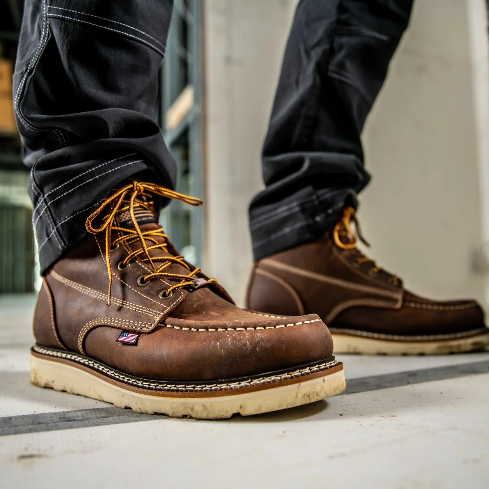 Close-up view of a person wearing brown leather work boots with tan soles and yellow laces, standing on a concrete surface. The boots feature detailed stitching and a small American flag patch on the side.