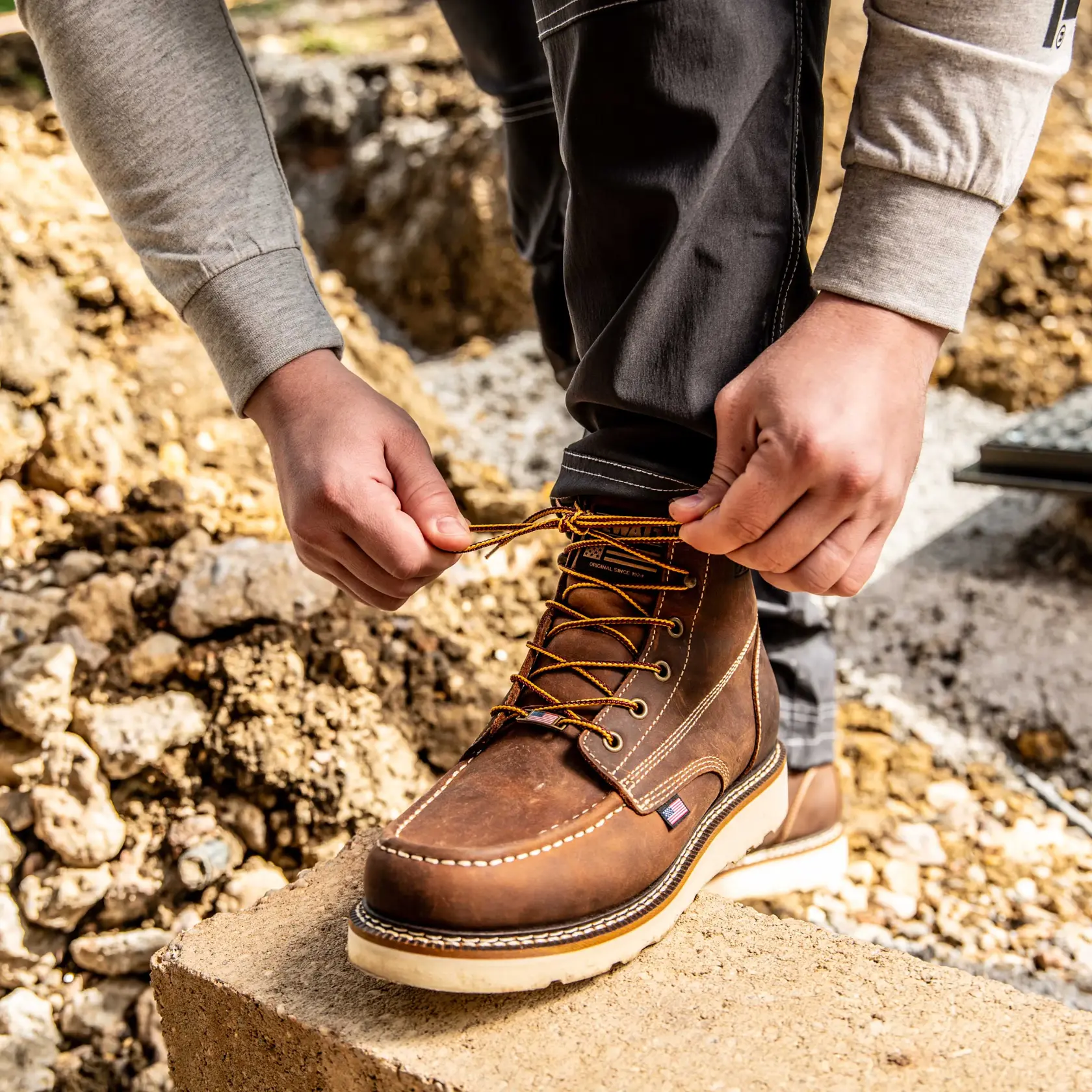 A person is tying the laces of a brown work boot while standing outdoors on a concrete block, with dirt and rocks in the background.
