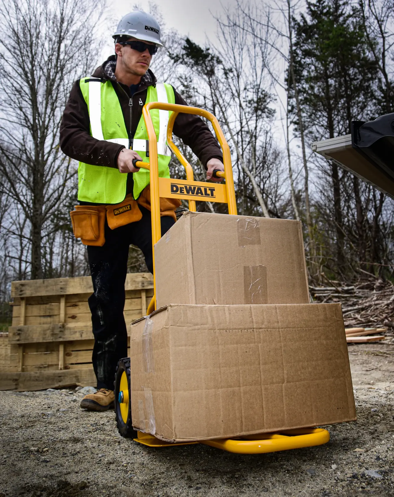 A construction worker wearing a safety vest and helmet uses a yellow DEWALT hand truck to transport two large cardboard boxes outdoors on a worksite.