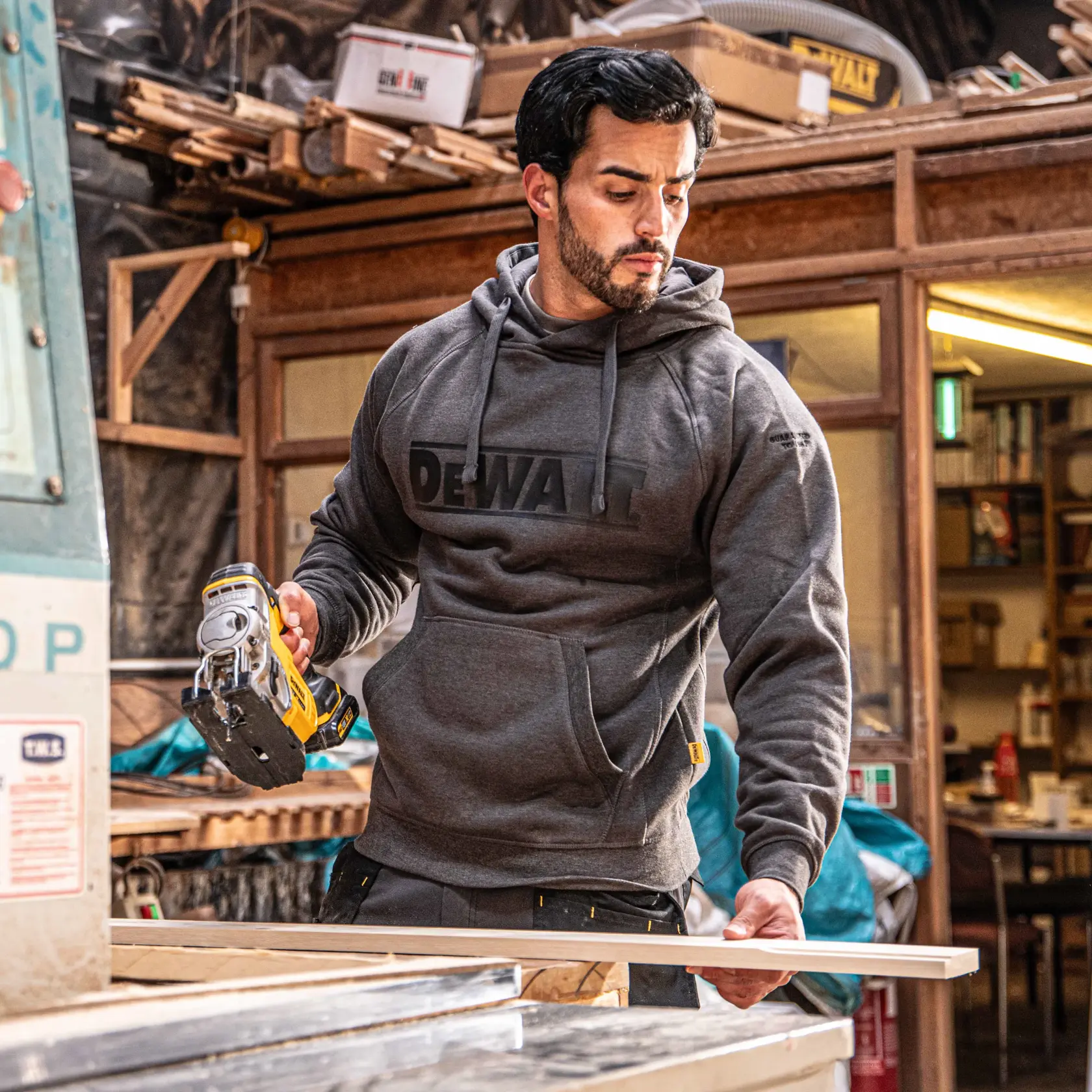 A person wearing a gray DEWALT hoodie is working in a woodworking shop, holding a yellow DEWALT power tool and a piece of wood. The face is blurred, and the workspace is filled with various tools and equipment.
