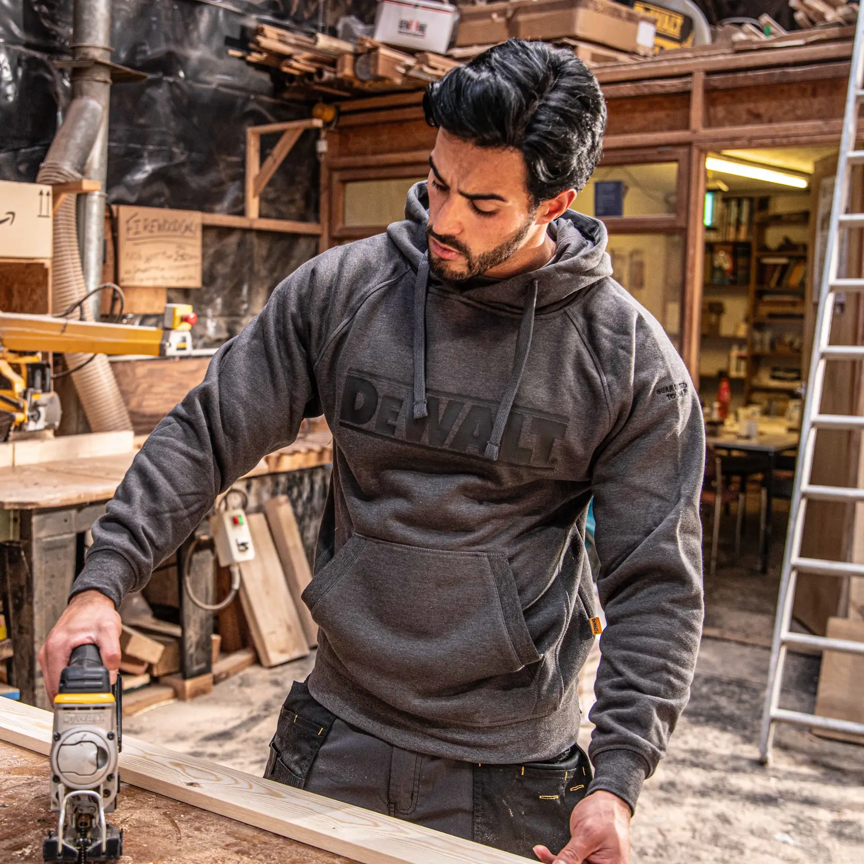 A person wearing a grey DEWALT hoodie is operating a woodworking tool on a piece of wood in a workshop. Their face is blurred for privacy.