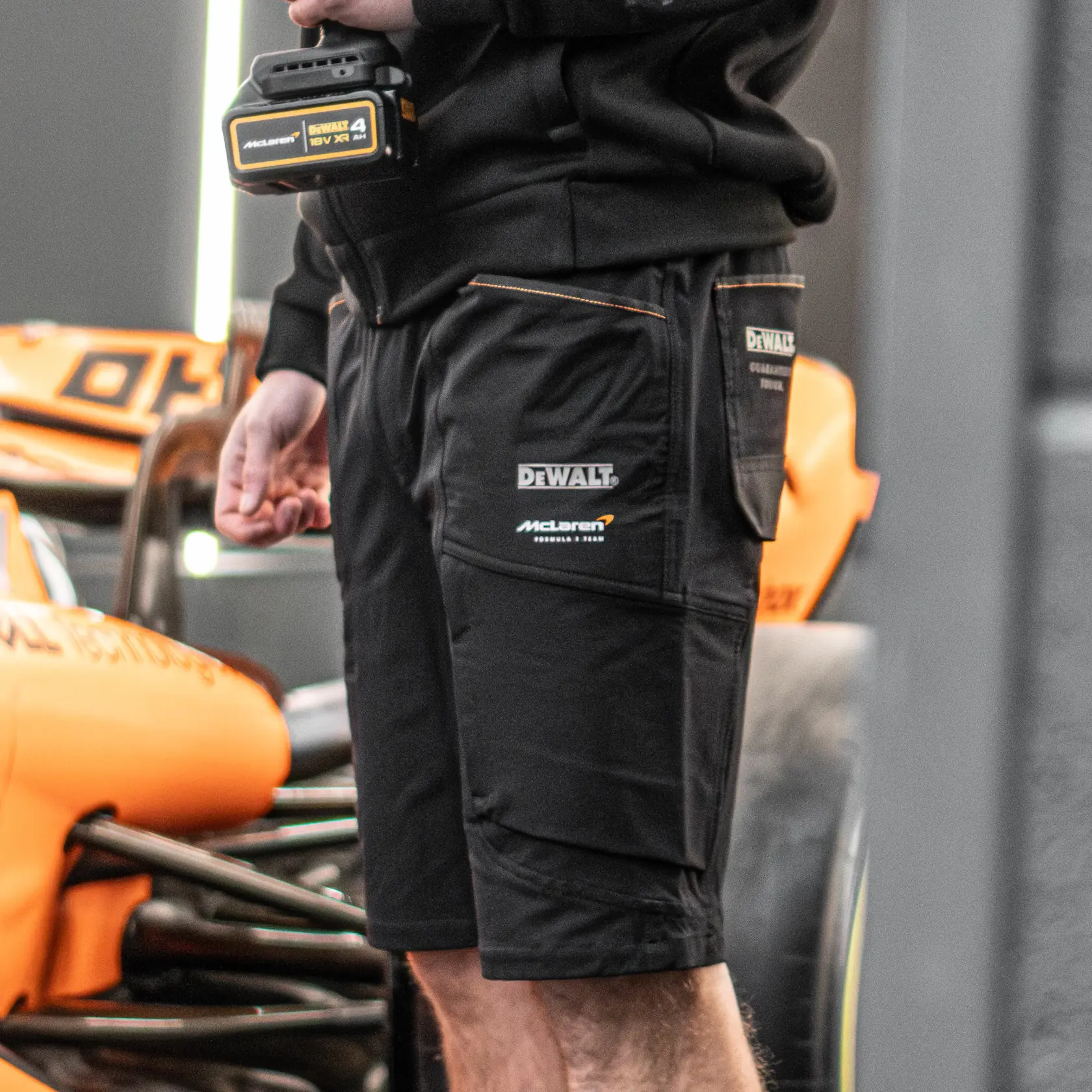 A person wearing black DEWALT and McLaren branded work shorts stands next to an orange vehicle part. The person is holding a DEWALT 18V XR battery, and detailed branding is visible on the shorts and battery.