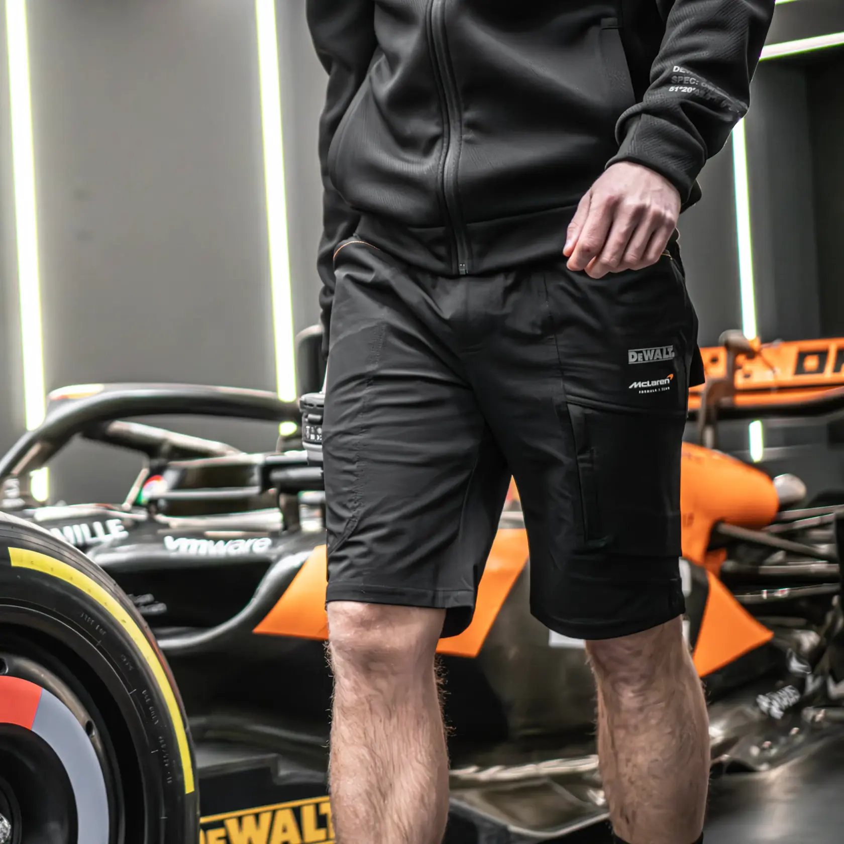 A person wearing a black DEWALT jacket and black DEWALT cargo shorts stands in front of a race car with black and orange detailing. The focus is on the lower half of the person's body and the DEWALT branding on the shorts.