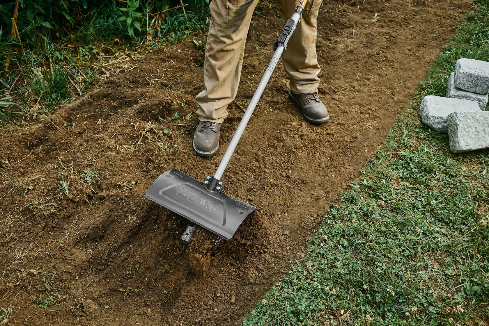 Person uses a DEWALT tool attachment to move dirt along a garden path bordered by grass and stones.