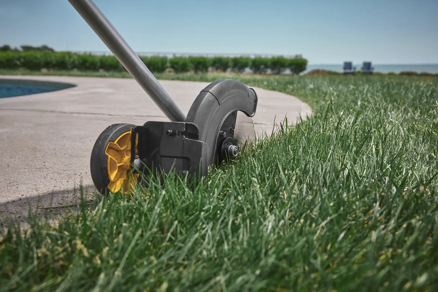 Lawn edger trimming grass along a concrete edge near a pool in a landscaped outdoor area.
