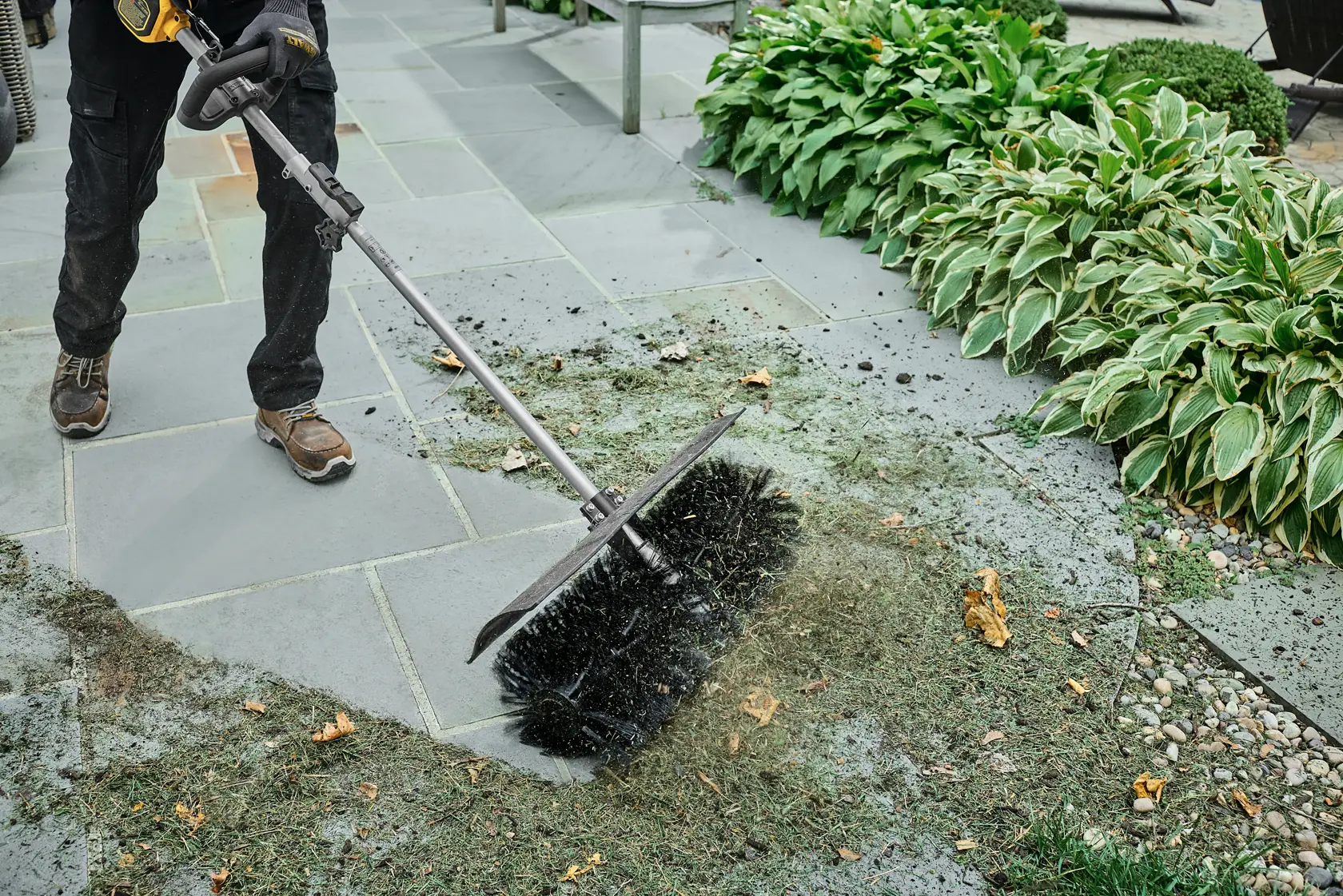 Person uses a DEWALT power broom attachment to clean grass and debris from outdoor stone patio near plants.