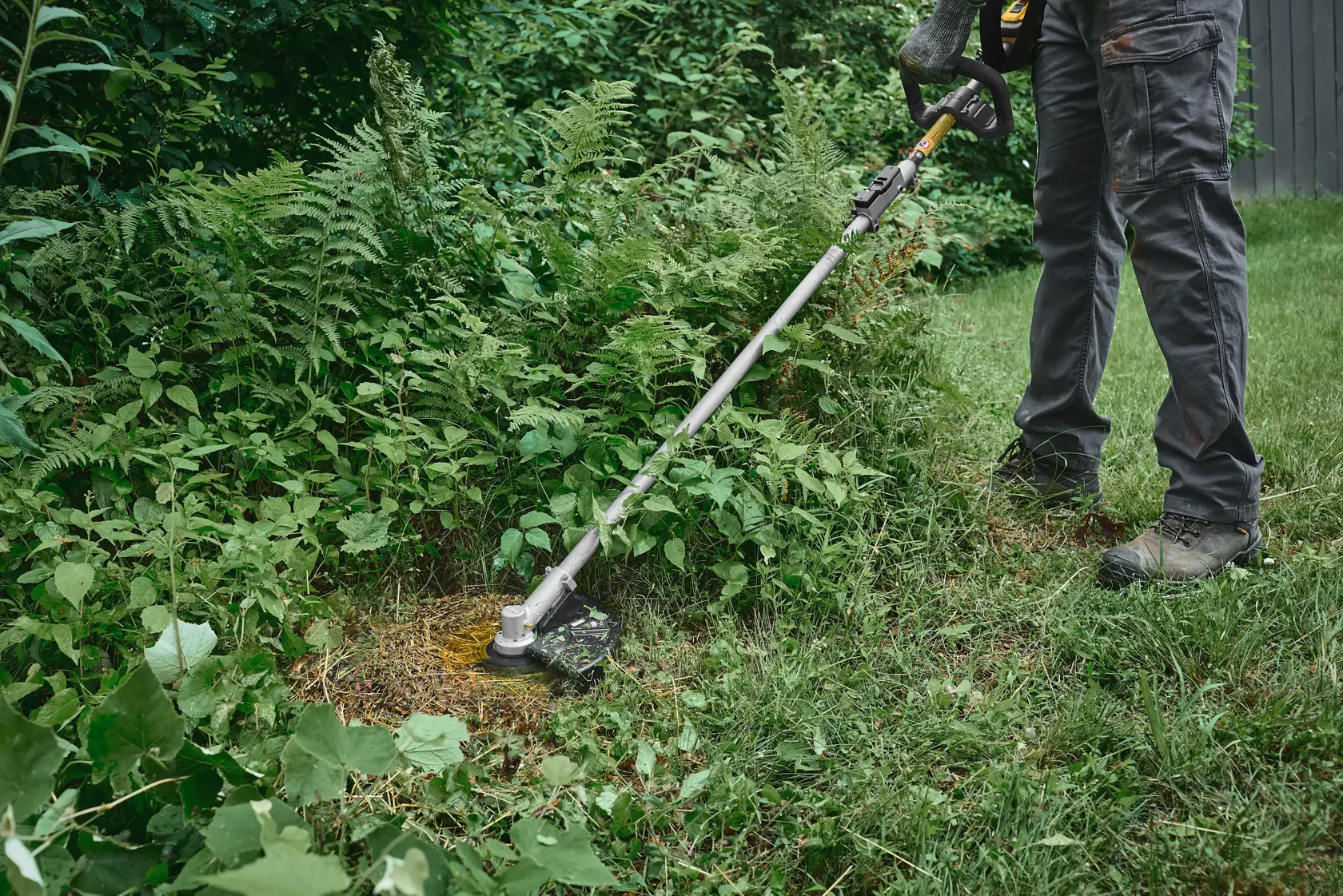 Person using DEWALT brush cutter to trim overgrown grass and weeds outdoors.