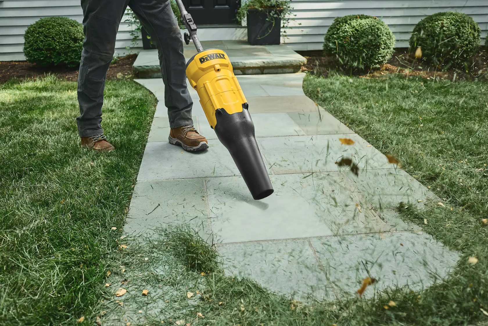 Person using a DEWALT leaf blower to clear grass and leaves from a stone path in a garden.