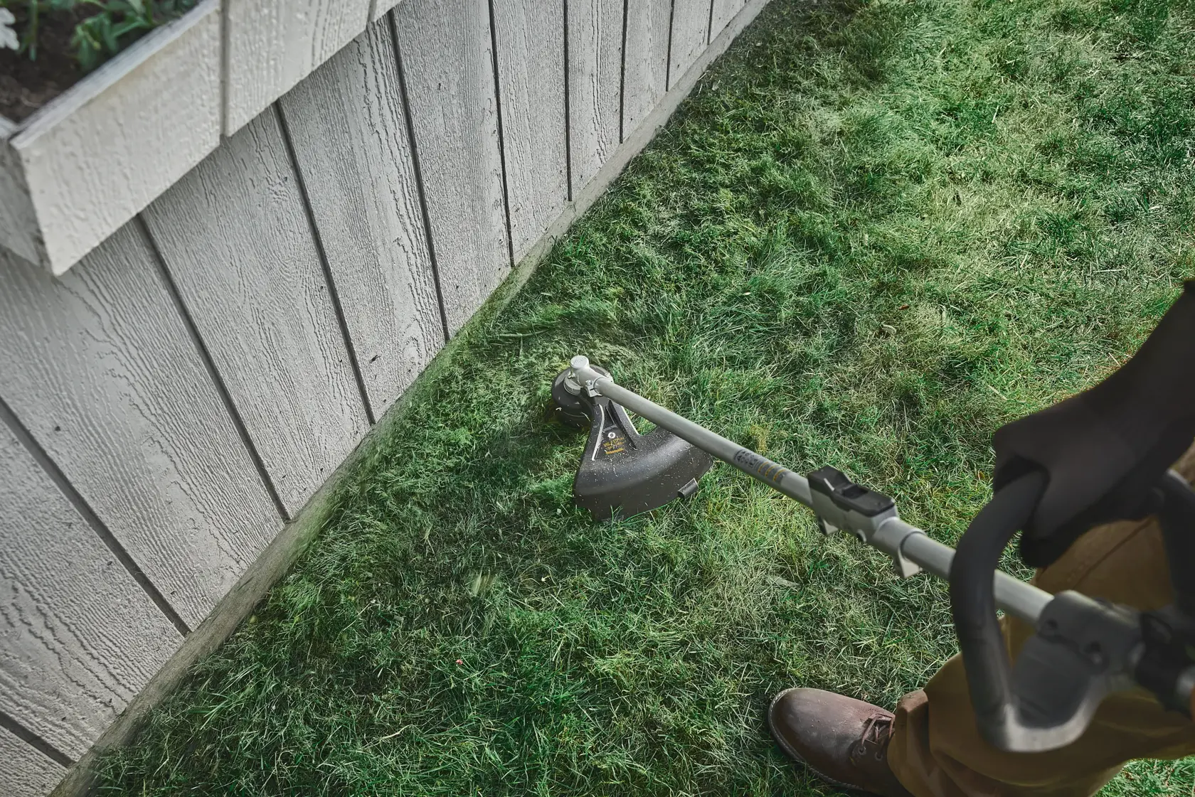 Person using a string trimmer to cut grass along the edge of a wooden building.