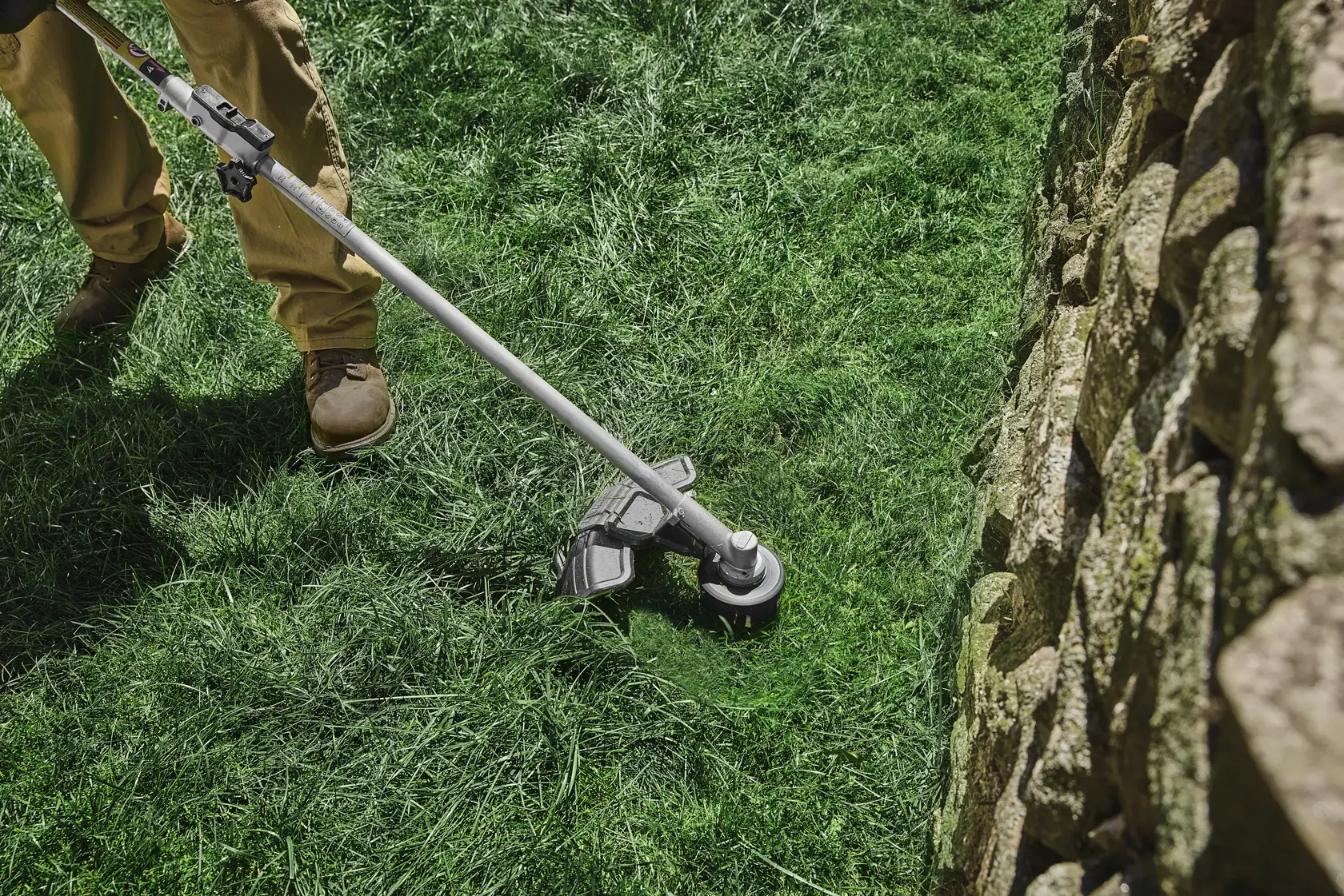 Person uses a DEWALT string trimmer to cut grass near a stone wall outdoors.