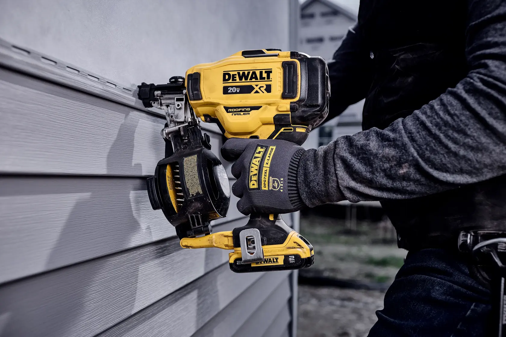A person wearing gloves uses a DEWALT 20V XR cordless roofing nailer to fasten siding on a wall. The tool is yellow and black and features the DEWALT logo prominently.